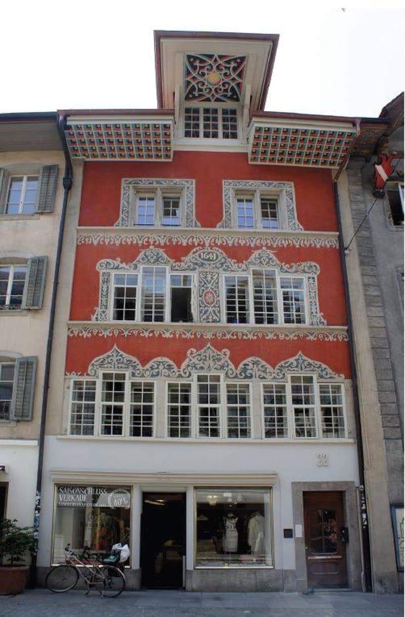 A historic red building with ornate white decorative trim around the windows and roofline. The ground floor features a storefront with large glass windows displaying clothing and accessories.