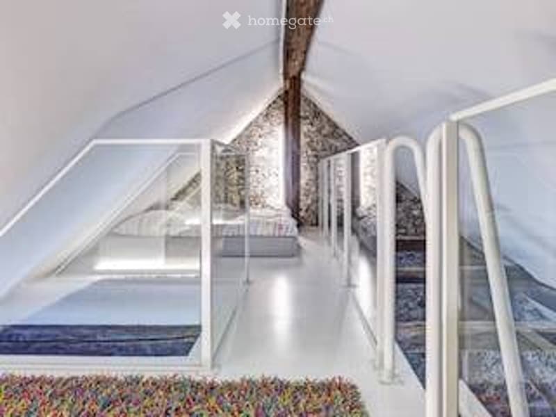 Bright, minimalist attic bedroom with white walls, exposed wooden beams, and a single bed on a raised platform. The room features large windows and a modern, airy design.