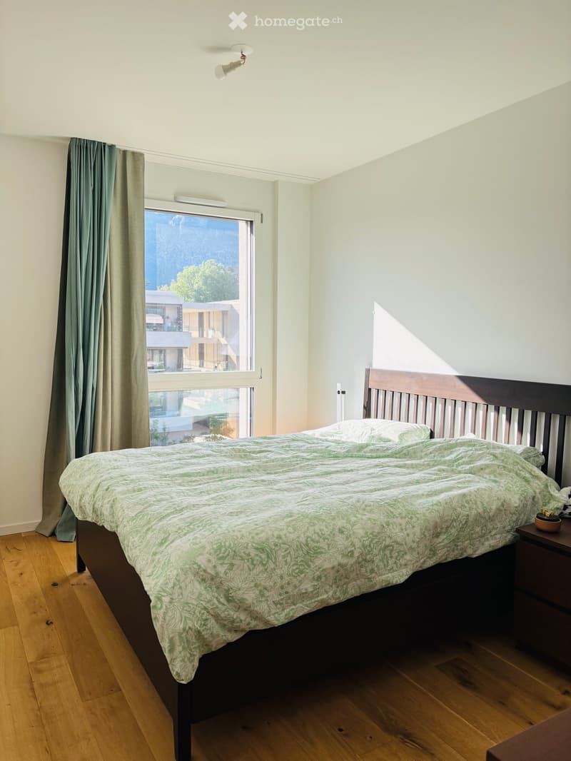 A cozy bedroom with a wooden bed, green patterned duvet, and a nightstand with a small bowl. The room features wooden flooring, white walls, and a large window with green curtains offering a view of the outside.