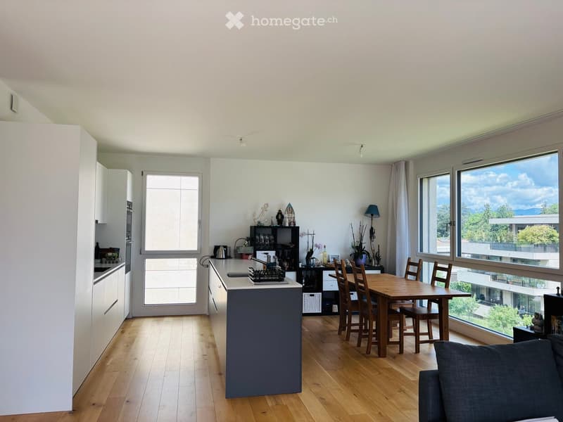 Open-plan living and dining area with a kitchen island, dining table, chairs, and a sofa, featuring large windows with a scenic view.