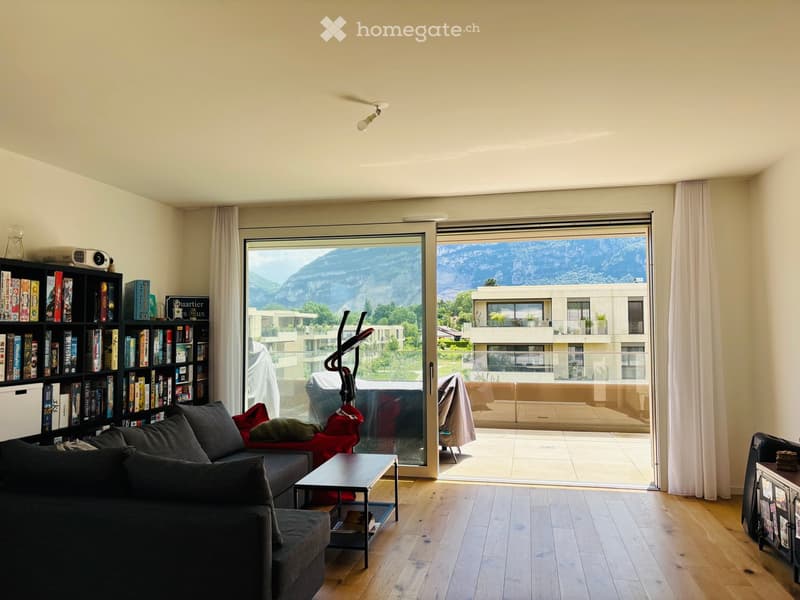 Modern living room with large windows offering a scenic mountain view, featuring a grey sectional sofa, a black coffee table, a bookshelf filled with books, and an exercise bike in the corner.