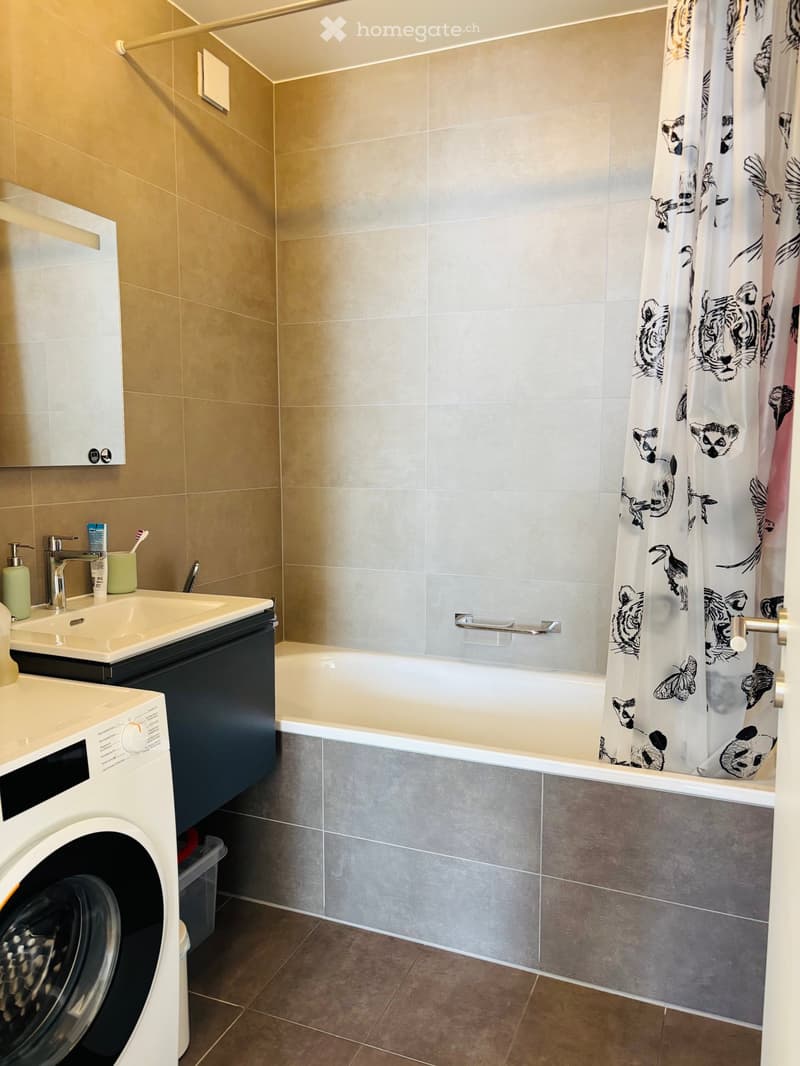 Modern bathroom with a bathtub, sink, mirror, and washing machine. The walls are tiled in beige and grey, and there is a decorative shower curtain with animal prints.