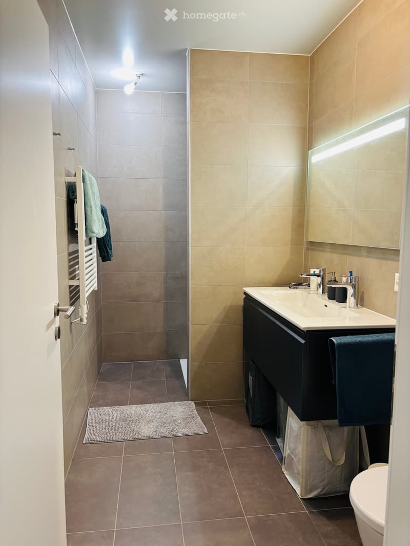 Modern bathroom with beige tiles, a white sink with a black cabinet, a large mirror, a toilet, a towel rack with towels, and a small rug on the floor.