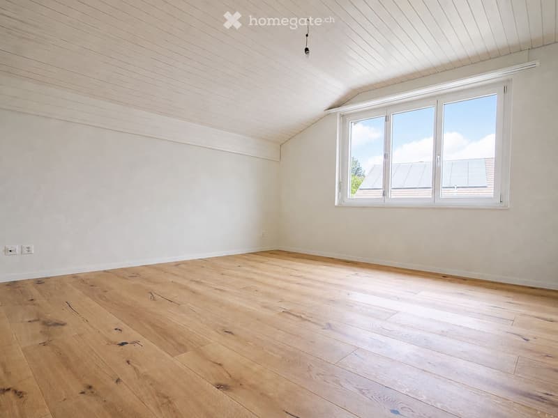 Empty room with wooden flooring, white walls, and a large window with white frame. The ceiling has a slanted design with wooden paneling.