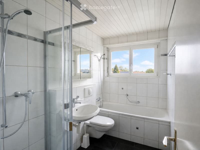 Modern bathroom with white tiles, a bathtub, toilet, sink, and a glass-enclosed shower area. Large windows provide natural light.