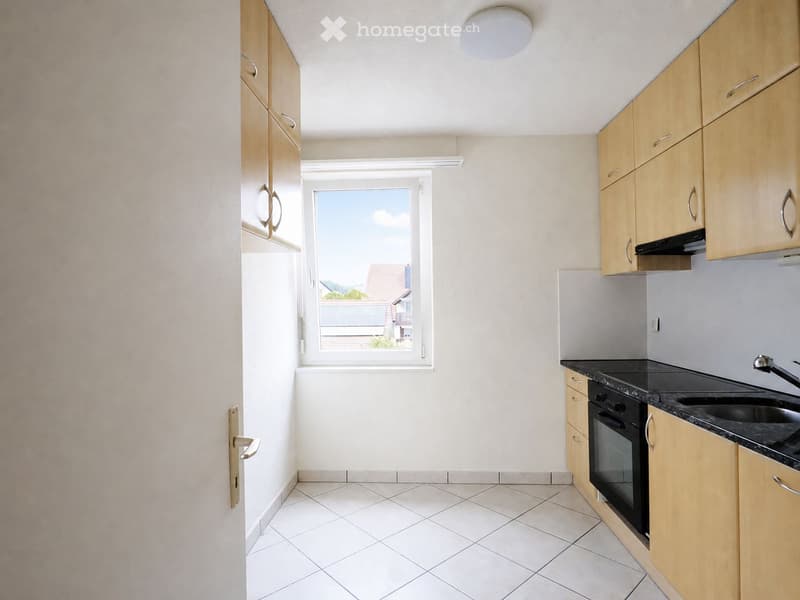 Modern kitchen with light wooden cabinets, black countertop, sink, and oven. White tiled floor and large window offering a view of the outside.