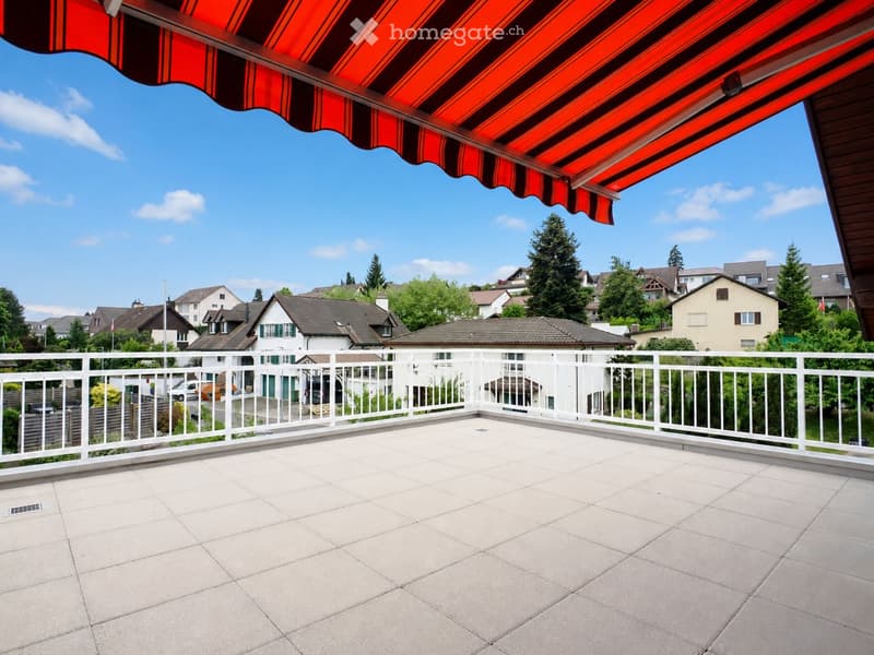 Balcony with tiled floor, white railing, and red and black striped awning, overlooking a residential neighborhood with houses and trees.