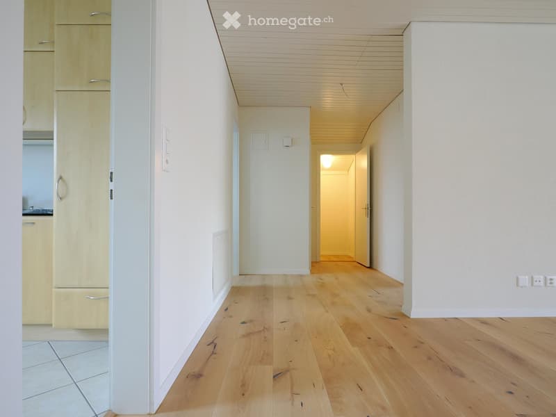 Empty hallway with light wooden flooring, white walls, and a visible kitchen area on the left side.