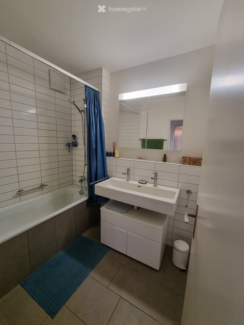 Modern bathroom with a bathtub, sink, mirror, toilet, and a blue bath mat on the floor.