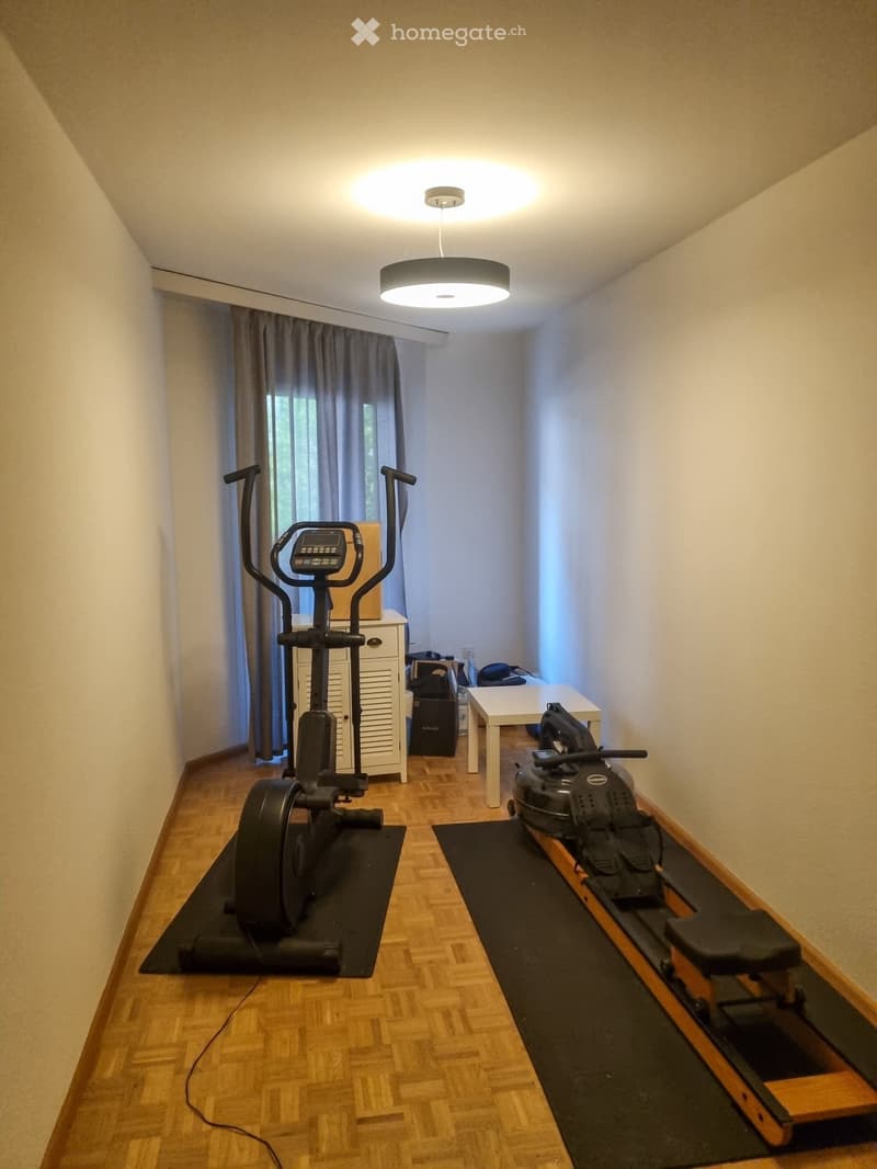 Well-lit room with wooden flooring, exercise bike, elliptical machine, small table, and cabinet.