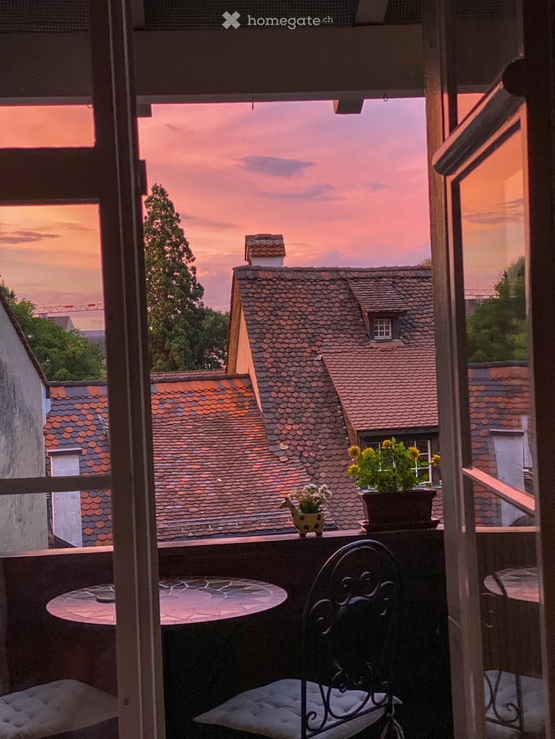 Balcony with a round table, cushioned chair, and potted plants, overlooking a cityscape at sunset.