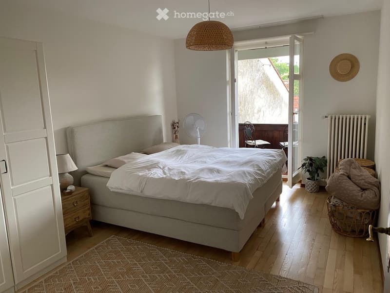 Cozy bedroom with a large bed, nightstand, lamp, fan, chair, plant, wicker basket, radiator, and rug. Large window with open shutters lets in natural light.