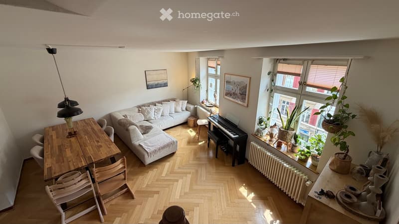A modern living room with a white sectional sofa, wooden dining table, chairs, piano, and several potted plants near the windows.