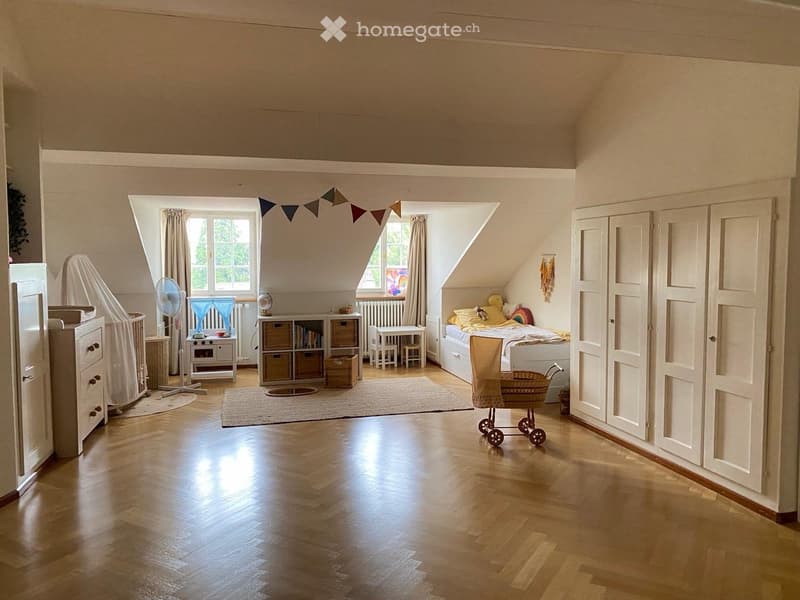 Bright, spacious bedroom with wooden floors, large windows, a bed with pillows and blankets, a wardrobe, a fan, a small table and chairs, storage units, and a baby cot.