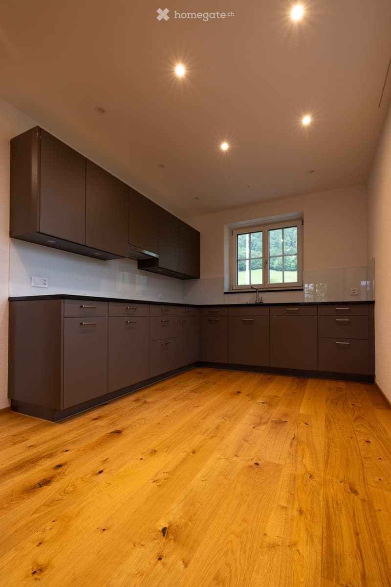 Modern kitchen with grey cabinets, wooden flooring, and a large window overlooking greenery.