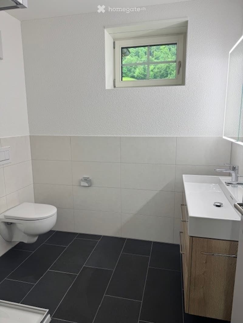 Modern bathroom with white tiled walls, black tiled floor, a toilet, a sink with a chrome faucet, and a window with a view of greenery.