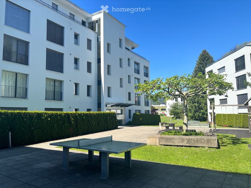 Courtyard with a ping pong table, benches, and a tree, surrounded by modern white buildings with balconies and large windows.