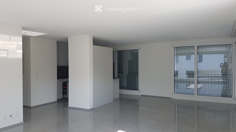 Empty room with white walls, dark tiled floor, large sliding glass doors, and a small kitchen area with cabinets and appliances.