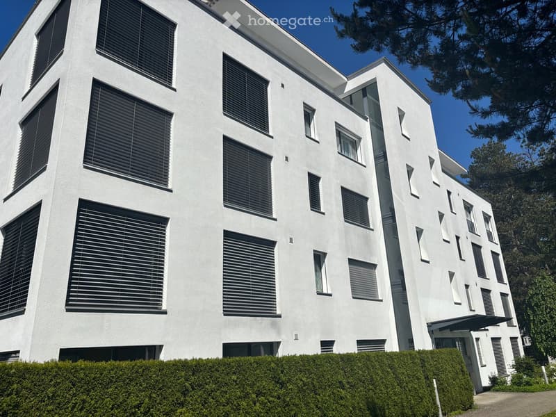 Modern white multi-story building with large windows and black blinds, surrounded by greenery and a neatly trimmed hedge.