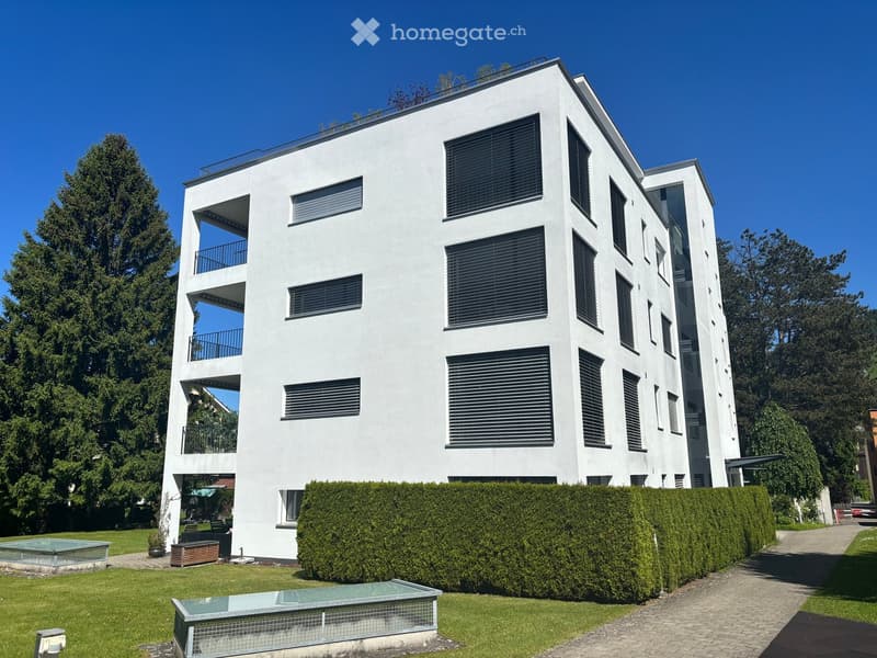 Modern white multi-story building with large windows and balconies, surrounded by a well-maintained garden with hedges, a small pool, and outdoor seating.