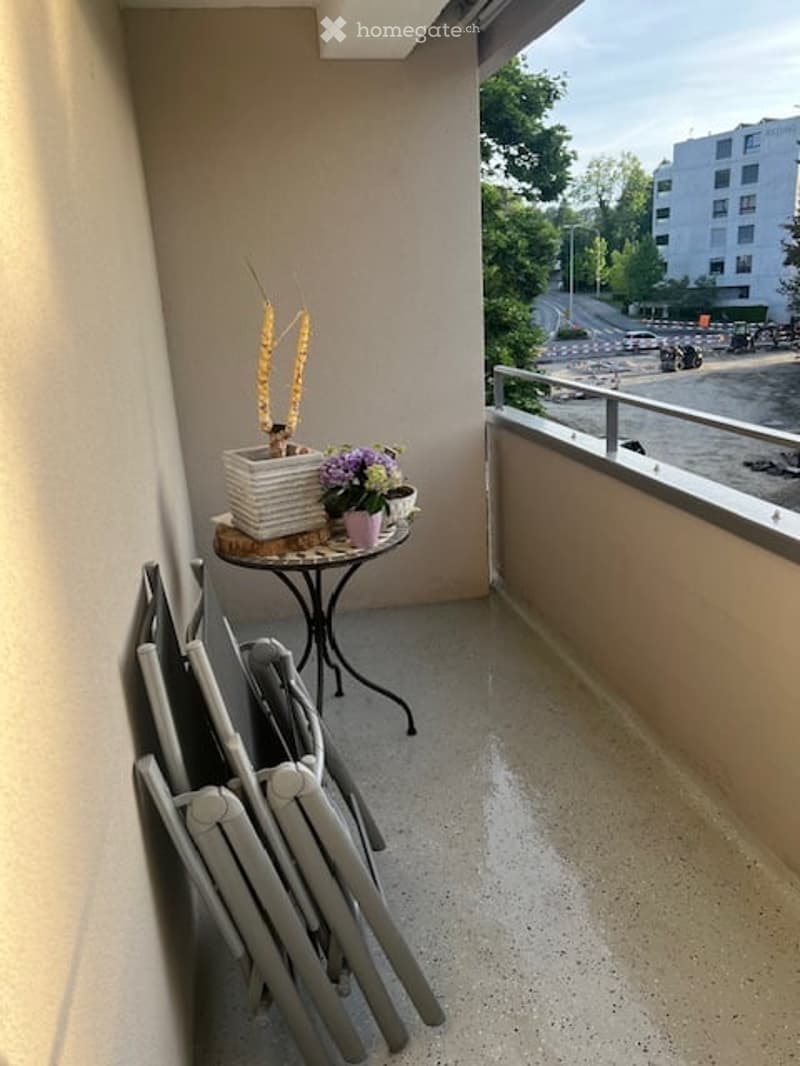 Balcony with a small table, potted plants, and stacked chairs, overlooking a street with buildings and greenery.