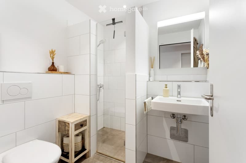 Modern bathroom with white tiles, a toilet, sink, mirror, and shower area. Includes a small wooden stool with a basket and decorative items on the shelves.