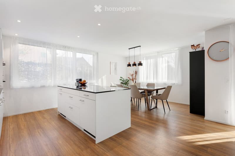 Bright, modern kitchen with white cabinets, a black countertop, a dining area with a glass table and chairs, pendant lights, and a large window with sheer curtains.