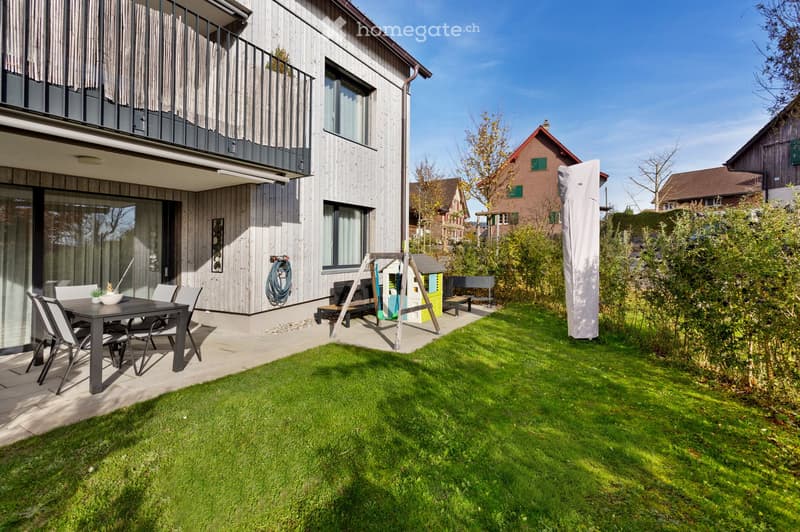 Well-maintained garden with a dining set, playhouse, and bench near a modern house with a balcony and large windows.