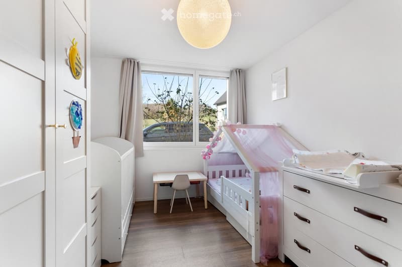 A well-lit, modern nursery with a crib, changing table, small desk, and chair. The room features a large window with curtains, a round ceiling light, and decorative elements like balloons and a picture frame.