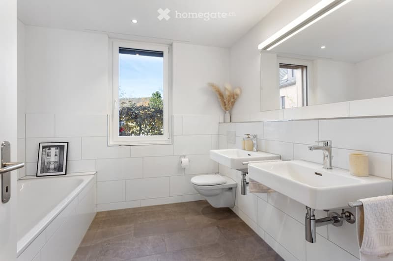 Modern bathroom with white tiles, double sinks, bathtub, toilet, large mirror, and decorative vase.
