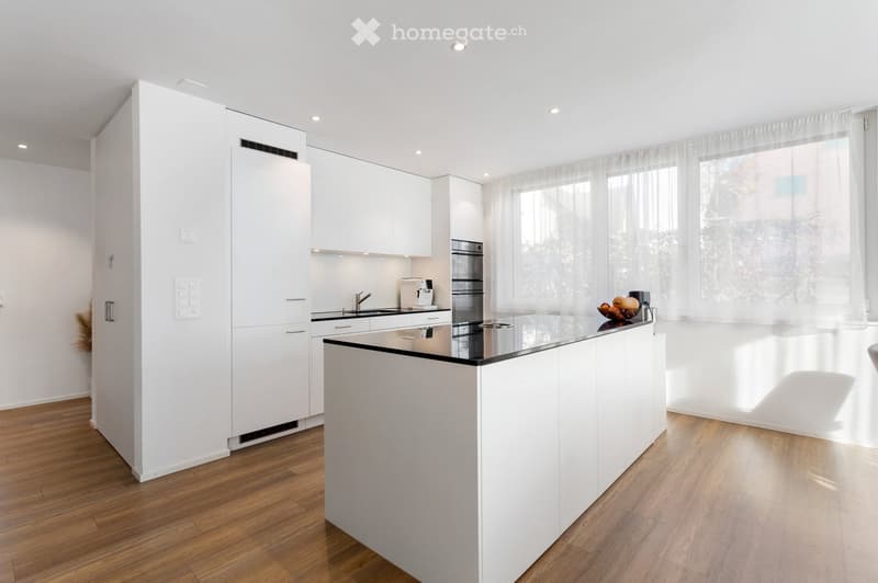Modern kitchen with white cabinets, a central island, and a black countertop. Features include a sink, faucet, coffee maker, and a refrigerator. Natural light streams in through large windows with sheer curtains.