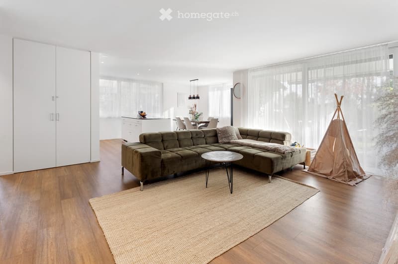 Modern living room with a green sectional sofa, coffee table, woven rug, and a small kitchen area with a dining table and chairs. A teepee structure is visible near the window.
