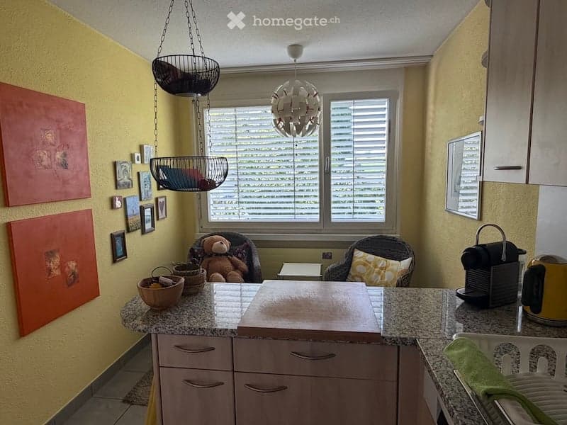 Bright kitchen with granite countertops, wooden cabinets, and a hanging wicker basket. Features a teddy bear in a wicker chair, a coffee maker, and several picture frames on the walls.