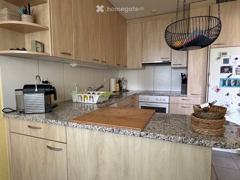 Modern kitchen with light wooden cabinets, granite countertops, and stainless steel appliances. Features include a coffee maker, cutting board, hanging wicker basket, and a sink with a faucet.