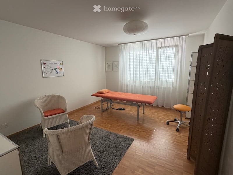 A small, well-lit room with wooden flooring, a massage table, two wicker chairs, a small stool, and a curtain covering a window. The room also features a picture frame on the wall and a grey rug on the floor.