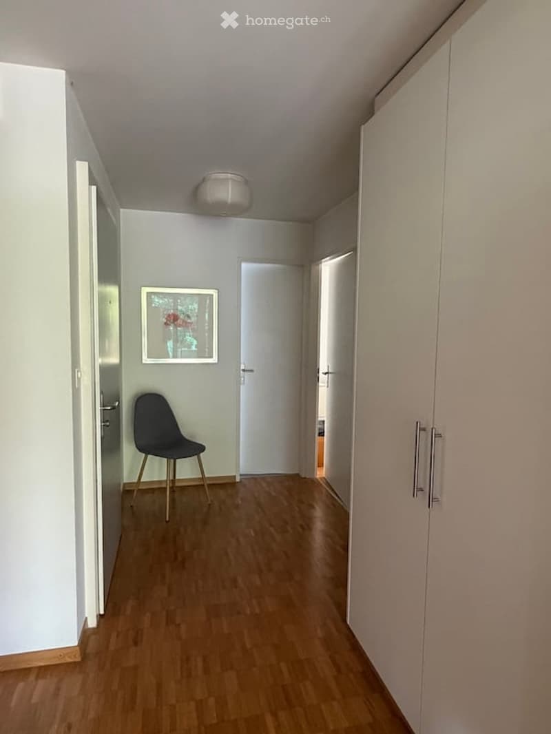 A narrow hallway with light grey walls, wooden flooring, and white cabinetry on both sides. A single chair is placed near the left wall, and a framed picture hangs above it. A door at the end of the hallway is slightly ajar, revealing another room.