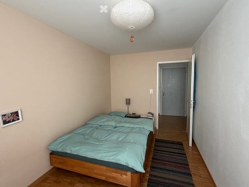 Cozy bedroom with a wooden bed, teal bedding, a nightstand with a lamp, and a small picture frame on the wall. The room has light-colored walls and a wooden floor.