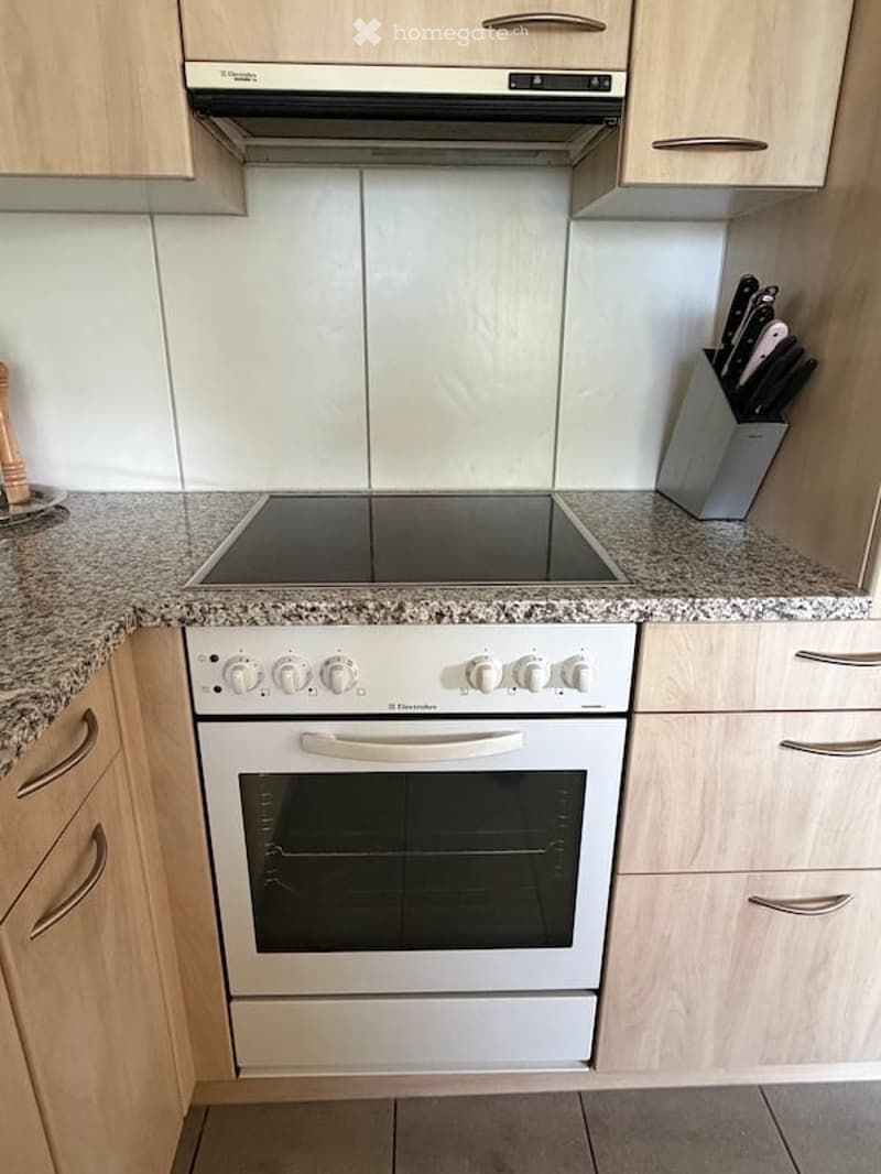 Modern kitchen with light wooden cabinets, granite countertops, an induction hob, and an electric oven. A knife block is visible on the counter.
