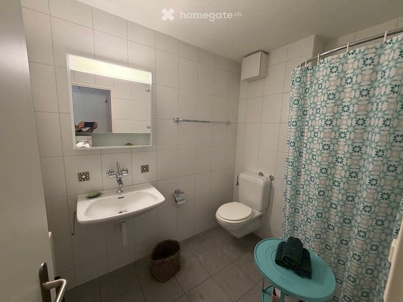 A small bathroom with white tiled walls and floor, featuring a sink, toilet, mirror, and a shower curtain. A small blue stool holds folded towels.