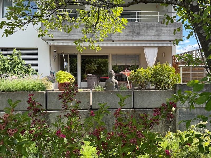 Garden area with a modern building in the background, featuring a balcony, potted plants, and a decorative statue.