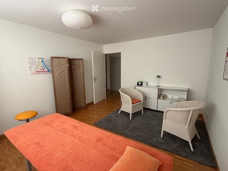 A small, well-lit room with a bed, two wicker chairs, a small orange stool, a cabinet, and a lamp. The room features wooden flooring and white walls.