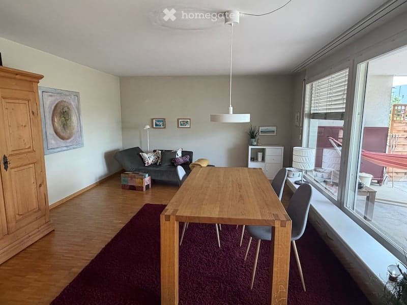A modern, well-lit living and dining area with a grey sofa, wooden dining table, and chairs. The space features a wooden cabinet, a floor lamp, and several picture frames on the wall. The room has large windows with blinds and a red carpet.