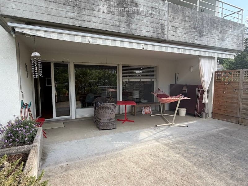 Balcony with outdoor furniture including a wicker chair, red table, and hammock, connected to a living space with large glass doors and white curtains.