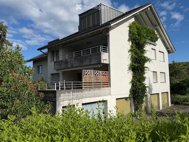 Modern two-story house with white exterior, multiple balconies, and greenery surrounding the property.
