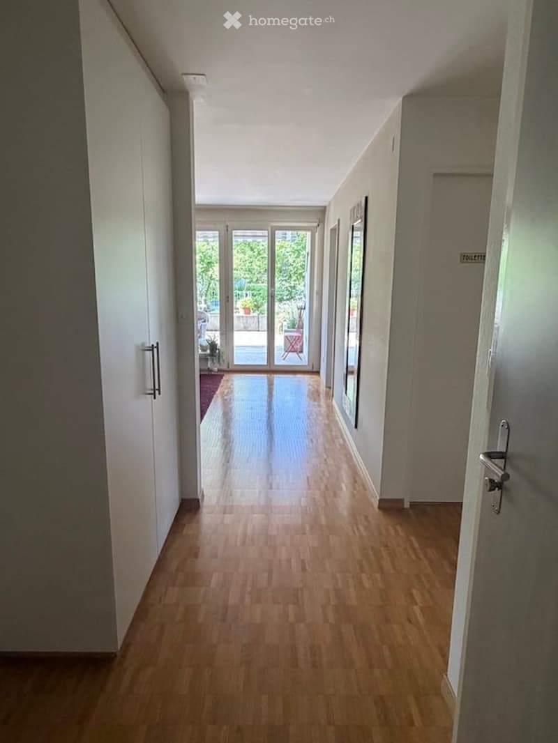 A long, well-lit hallway with wooden flooring and white walls, leading to a room with large glass doors and a view of a garden area.