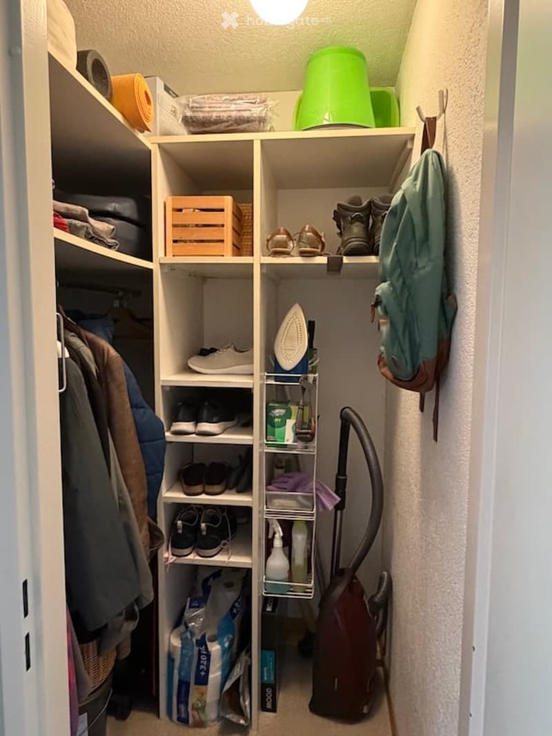A well-organized closet with multiple shelves containing clothes, shoes, a backpack, an ironing board, a vacuum cleaner, and cleaning supplies. The shelves are neatly arranged with various items including folded towels, a green bucket, and a wooden crate.