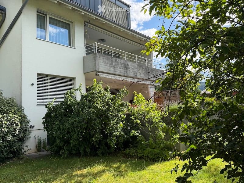Modern multi-story apartment building with white exterior, large windows, and a balcony on the upper floor. Surrounded by greenery and trees, with a clear view of the surrounding landscape.