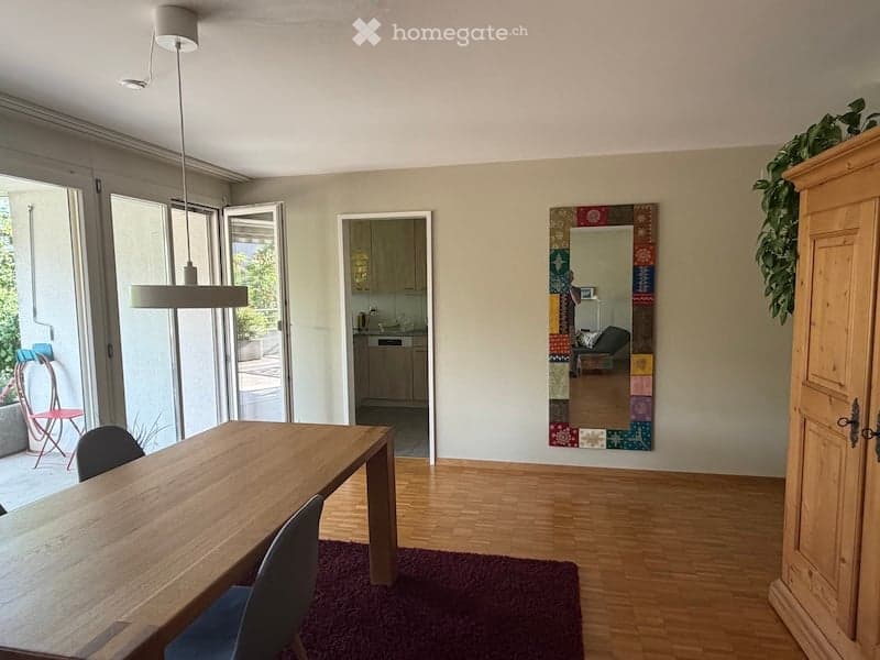 Bright, open-plan living and dining area with wooden flooring, large glass doors leading to a balcony, a dining table with chairs, a colorful mirror on the wall, and a cabinet.