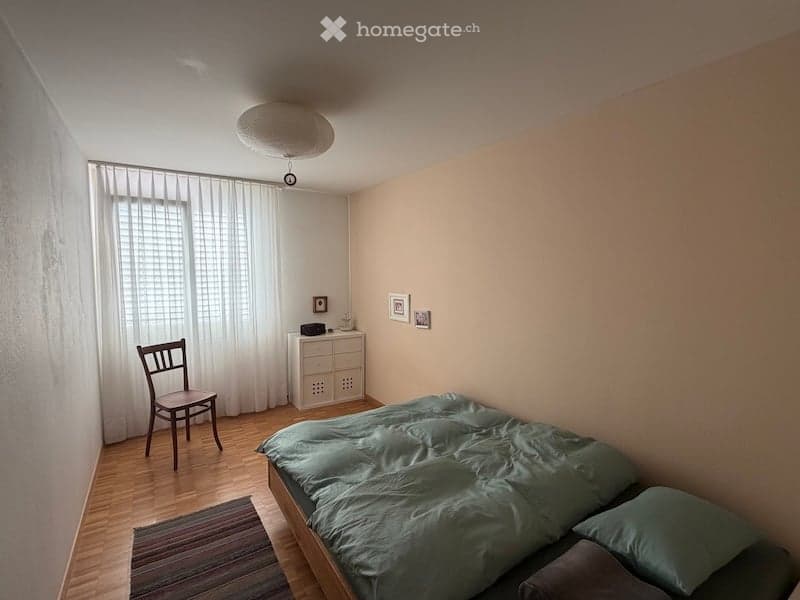 A small, cozy bedroom with a wooden bed, a wooden chair, a dresser, and a window with white curtains. The room features light-colored walls and a wooden floor.
