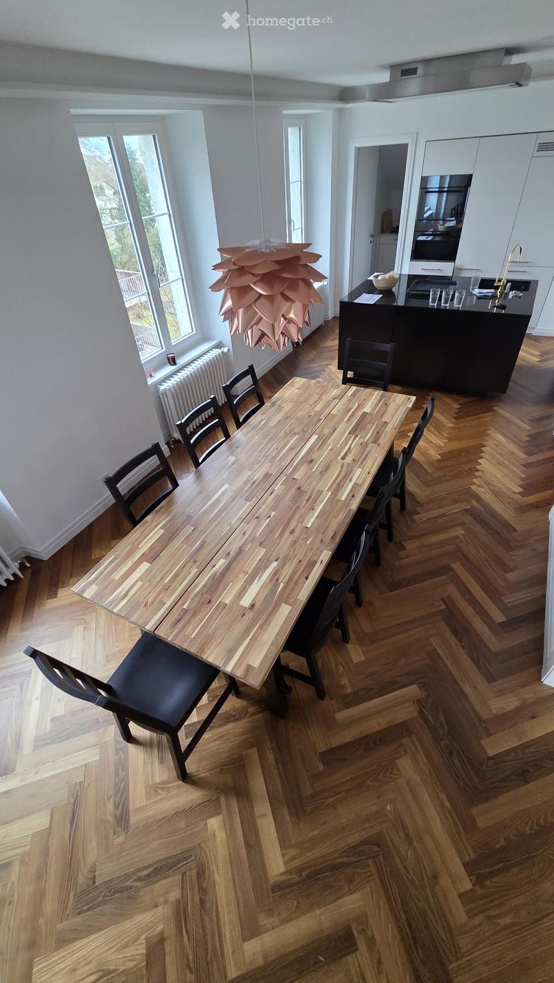 Modern kitchen with herringbone wooden flooring, a long wooden dining table surrounded by black chairs, a kitchen island with a sink, and a hanging light fixture.
